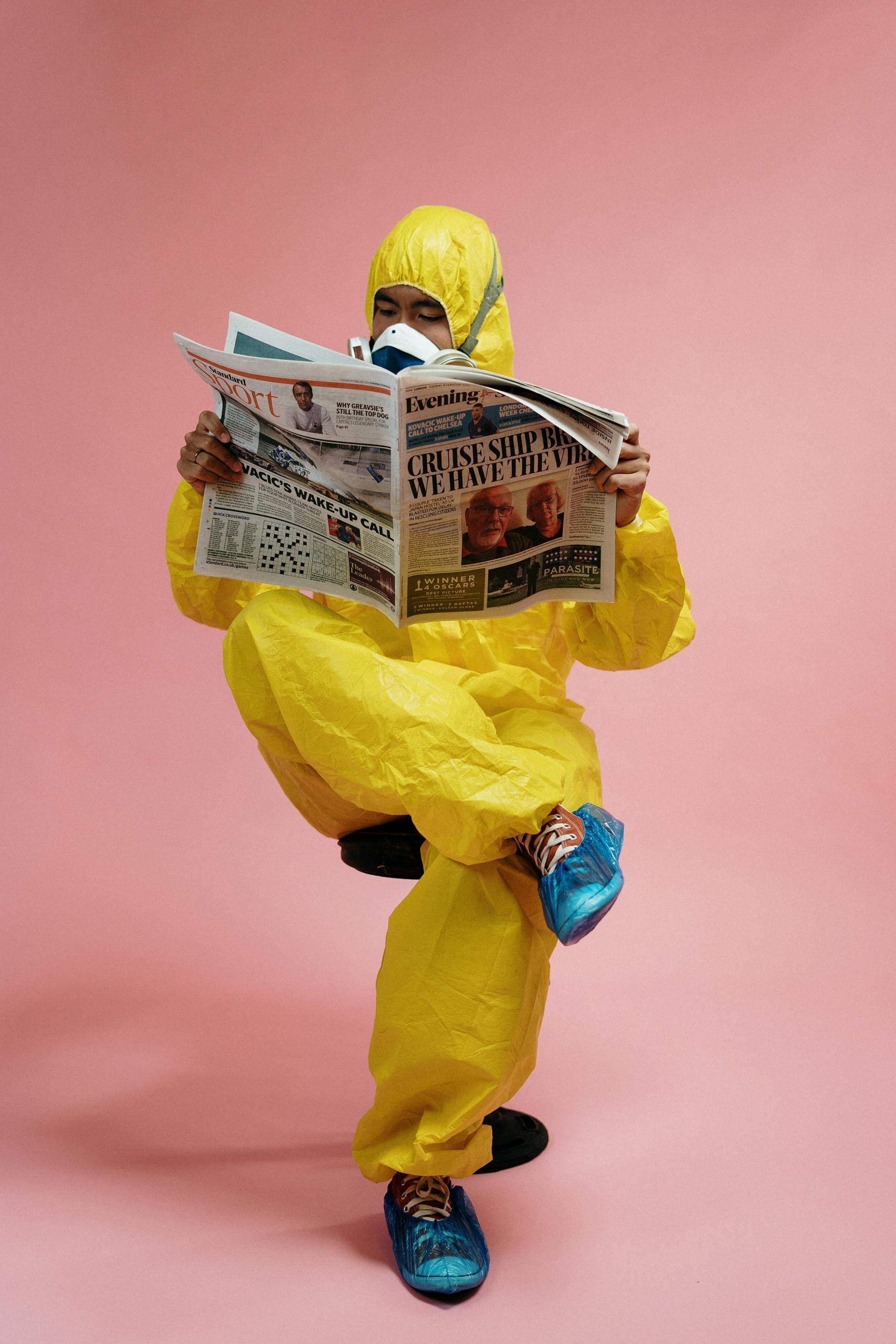 A man in a yellow hazmat suit and respirator reads a newspaper against a pink background.