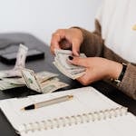 Close-up of person counting cash with notepad on desk, indicating financial tasks.