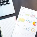 Flatlay of business report with colorful charts, a notebook, and a laptop for data analysis on a desk.
