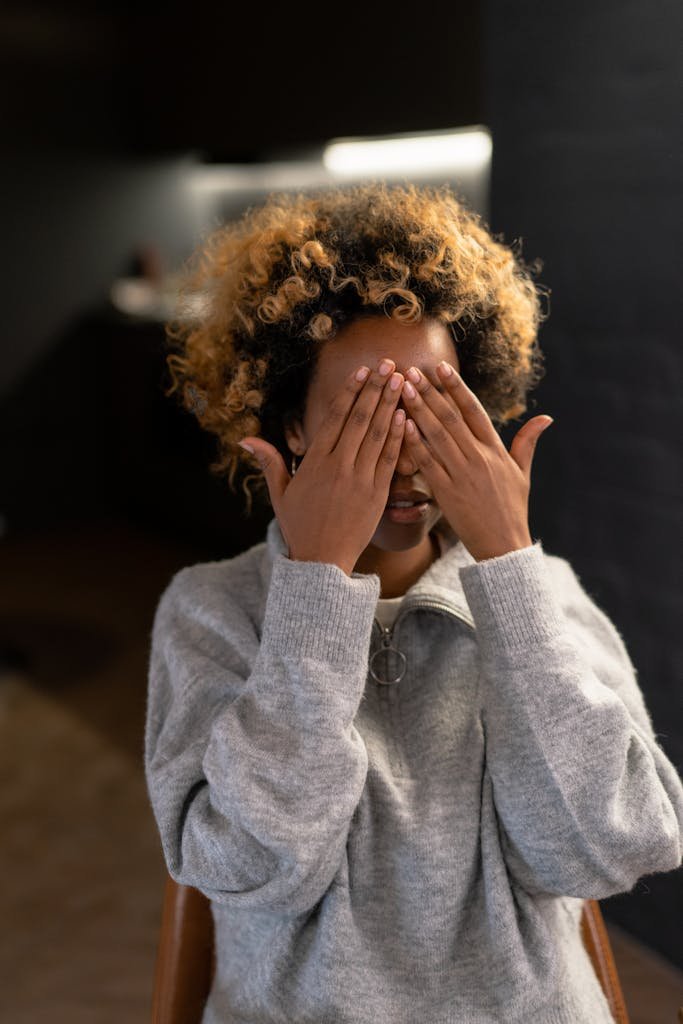 A woman with afro hair playfully covers her face with her hands indoors.