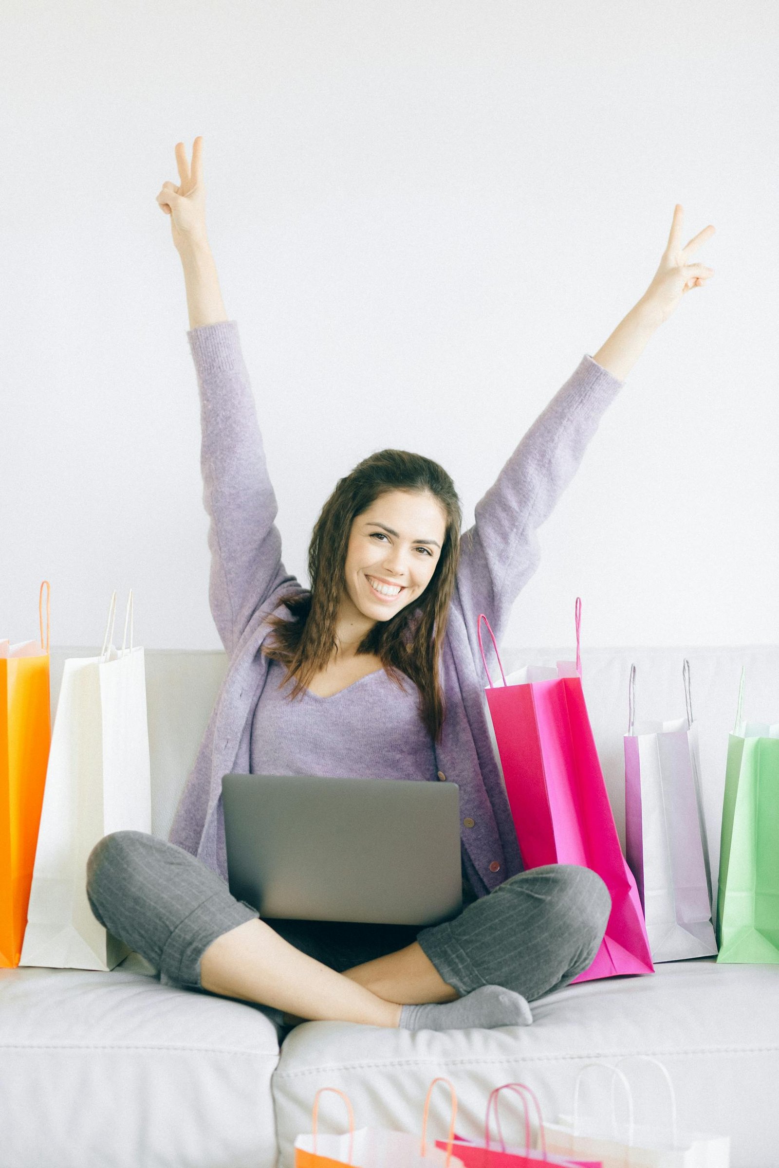 Cheerful woman sitting with laptop and shopping bags, celebrating online deals indoors.
