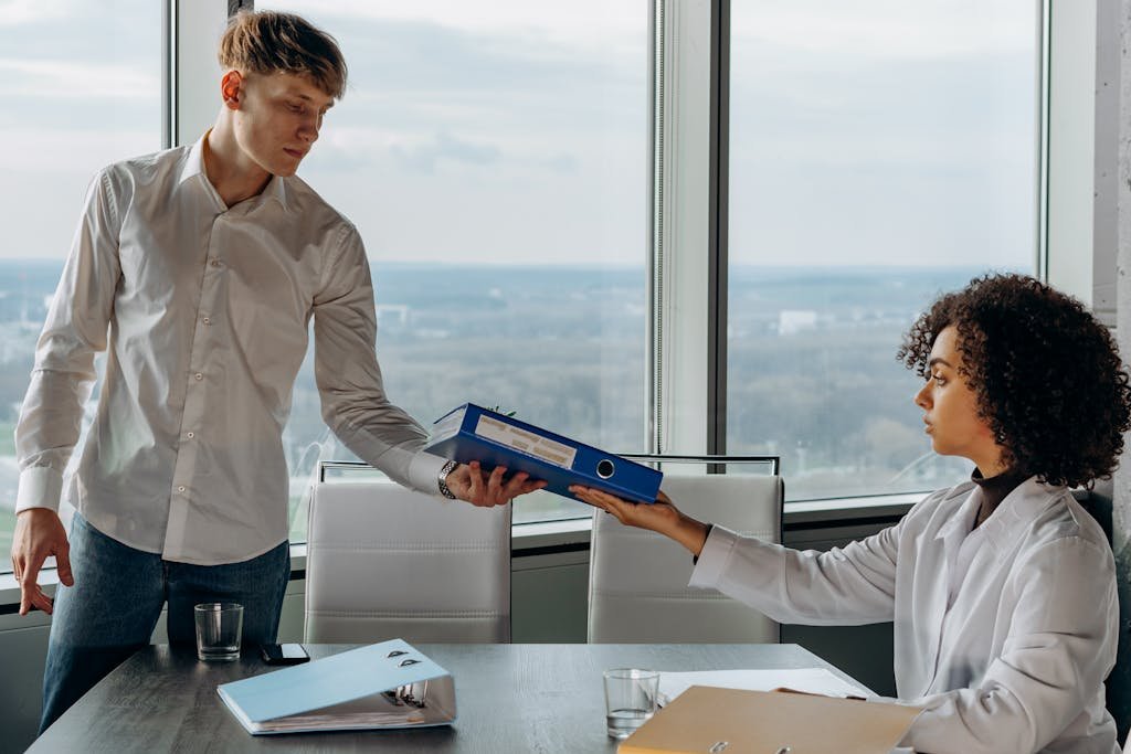 Two colleagues exchange documents in a contemporary office with a scenic city view.