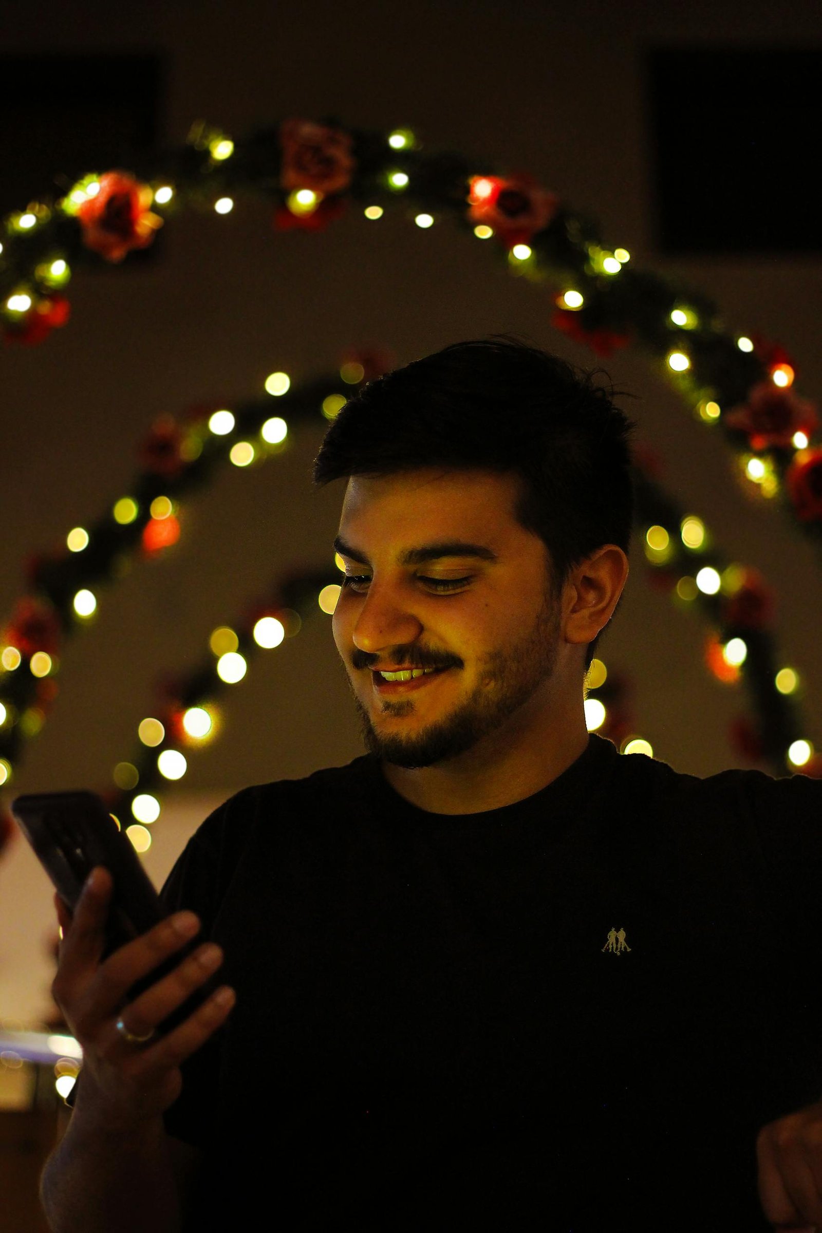 Young man smiling while using his smartphone under warm festive lights.